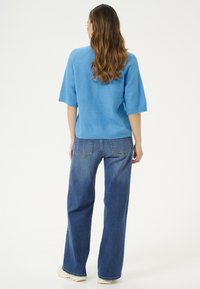 Femme aux longs cheveux portant un pull bleu à manches courtes, un jean bleu et des chaussures blanches, debout de dos sur un fond blanc.