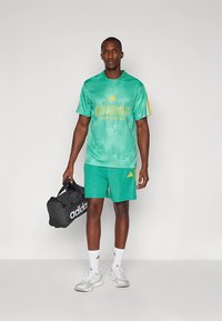 Πράσινο αθλητικό πουκάμισο με κίτρινες λεπτομέρειες και λογότυπο, συνδυασμένο με αντίστοιχα σορτς. Μαύρη σακούλα ταξιδιού με λευκή επωνυμία. Λευκά αθλητικά παπούτσια.