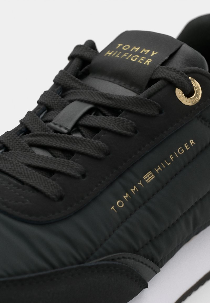 Czarne sneakerki z syntetycznej cholewki, pikowanym designem, złotym logo oraz płaskimi czarnymi sznurowadłami. Posiadają teksturowane akcenty i białą podeszwę.
