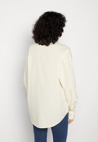 Chemise à manches longues en coton crème, avec un ourlet arrondi et des poignets boutonnés, présentée de dos.