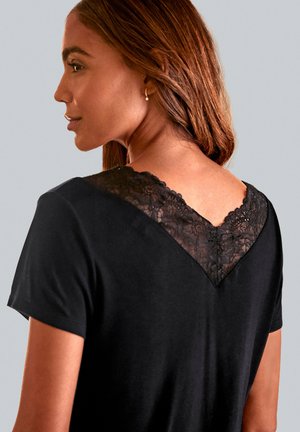 Eine Frau mit mittelbraunem Haar trägt ein schwarzes Oberteil mit einem Spitzen-V-Ausschnitt vor einem schlichten hellgrauen Hintergrund.