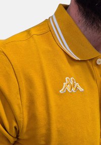 Polo giallo con superficie testurizzata, caratterizzato da un colletto a righe bianche e un logo ricamato sul petto.