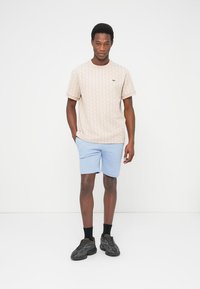 Maglietta beige a motivo con maniche corte, pantaloncini blu e sneakers nere, in piedi su uno sfondo neutro. Subile dettaglio logo sulla maglietta.