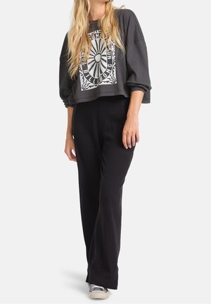 Kvinde iført sorte bukser med brede ben og en mørk cropped sweatshirt med hvidt motiv, stående i afslappet stilling, lette sneakers synlige.