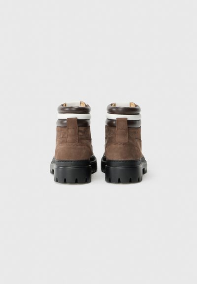 Une paire de bottines en daim marron avec des semelles en caoutchouc noir et des cols rembourrés blancs et marron foncé, vue de l'arrière sur un fond blanc.