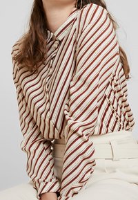 Long-sleeve blouse in cream with maroon stripes and thin accent lines. Features a buttoned collar and gathered detail at the front.