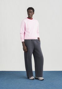 Sweatshirt rosa claro com punhos canelados e decote vermelho, combinado com calças de fato de treino cinza largas e sapatilhas cinzentas num fundo azul.