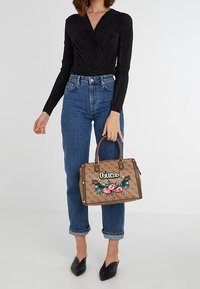Brun Guess håndtaske med broderet blomsterdesign, afrundede håndtag og guld logo. Båret med tætsiddende sort top og blå jeans.