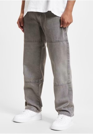 Man met losse grijze cargobroek en witte sneakers, staand tegen een effen lichte achtergrond.