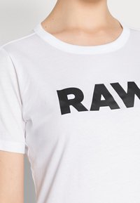 Bílý bavlněný tričko s kulatým výstřihem a krátkými rukávy. Na přední straně má velké černé písmo "RAW". Hladká textura.