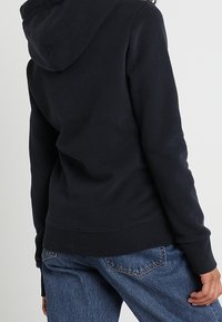 Sudadera oscura de color azul marino con una textura suave, que cuenta con capucha ajustable y puños y bajo de canalé. Lleva puesta con unos vaqueros de mezclilla azul claro.