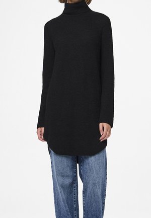 Osoba v dlouhém černém roláku s dlouhými rukávy a volných modrých džínách na bílém pozadí.