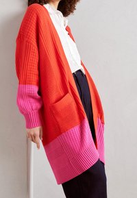 Osoba ubrana w czerwoną i różową teksturowaną dzianinową kurtkę na białej koszuli z guzikami i ciemnych spodniach, oparta o jasną ścianę.