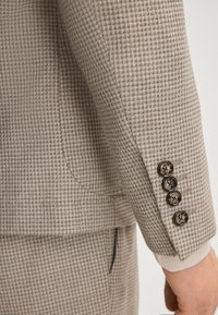 Rękaw marynarki w brązową kratkę z czterema ciemnymi guzikami, widoczna delikatna faktura, dopasowany krój, standardowy klap, oraz kieszeń boczna.