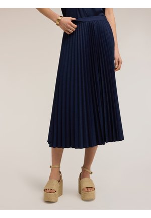 Kobieta ubrana w granatową plisowaną spódnicę midi, beżowe sandały na platformie z paskami przy kostce oraz złote bransoletki na jednym nadgarstku.