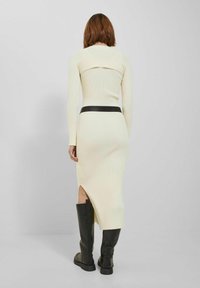 Kvinna med kort brunt hår, klädd i en krämfärgad ribbad långärmad midiklänning, svart bälte och knähöga svarta stövlar, stående med ryggen vänd.