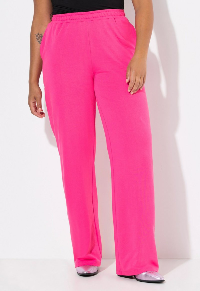 Un pantalon de jogging rose, léger et ample, avec une taille élastique et des poches latérales, associé à des chaussures brillantes en argent. Texture lisse.