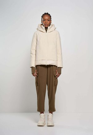 Donna con capelli intrecciati che indossa una giacca beige con cappuccio, pantaloni cargo marroni e stivali beige, in piedi davanti a uno sfondo chiaro e uniforme.