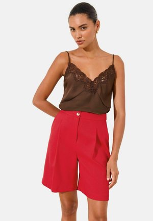 Frau trägt ein braunes Spitzen-Camisole, gesteckt in hoch taillierte, knallrote Shorts bis zum Knie vor einem schlichten Hintergrund.
