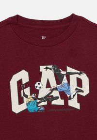 Bordó póló nagy "GAP" felirattal és két futballs játékost ábrázoló illusztrációval, az egyik csúszik a labda megkezdéséhez, a másik pedig ugrik, hogy elfogja azt.