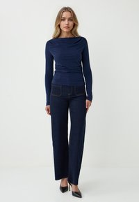 Maglia a maniche lunghe di colore navy con scollatura drappeggiata abbinata a pantaloni a gamba larga in denim scuro con cuciture a contrasto e tasche posteriori, completati da tacchi neri.