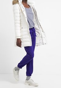 Bílá péřová bunda s kapucí lemovanou kožešinou, světle šedá košile, fialové tepláky, světle šedé ponožky a bílé boty ve stylu brogue.