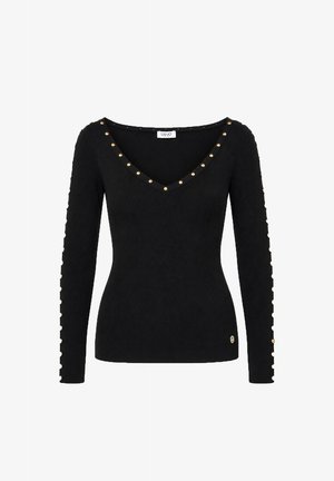 Mujer con una camiseta negra de manga larga con tachuelas doradas y pantalones beige de pierna ancha, de pie con una mano en el bolsillo y botas negras de tacón.