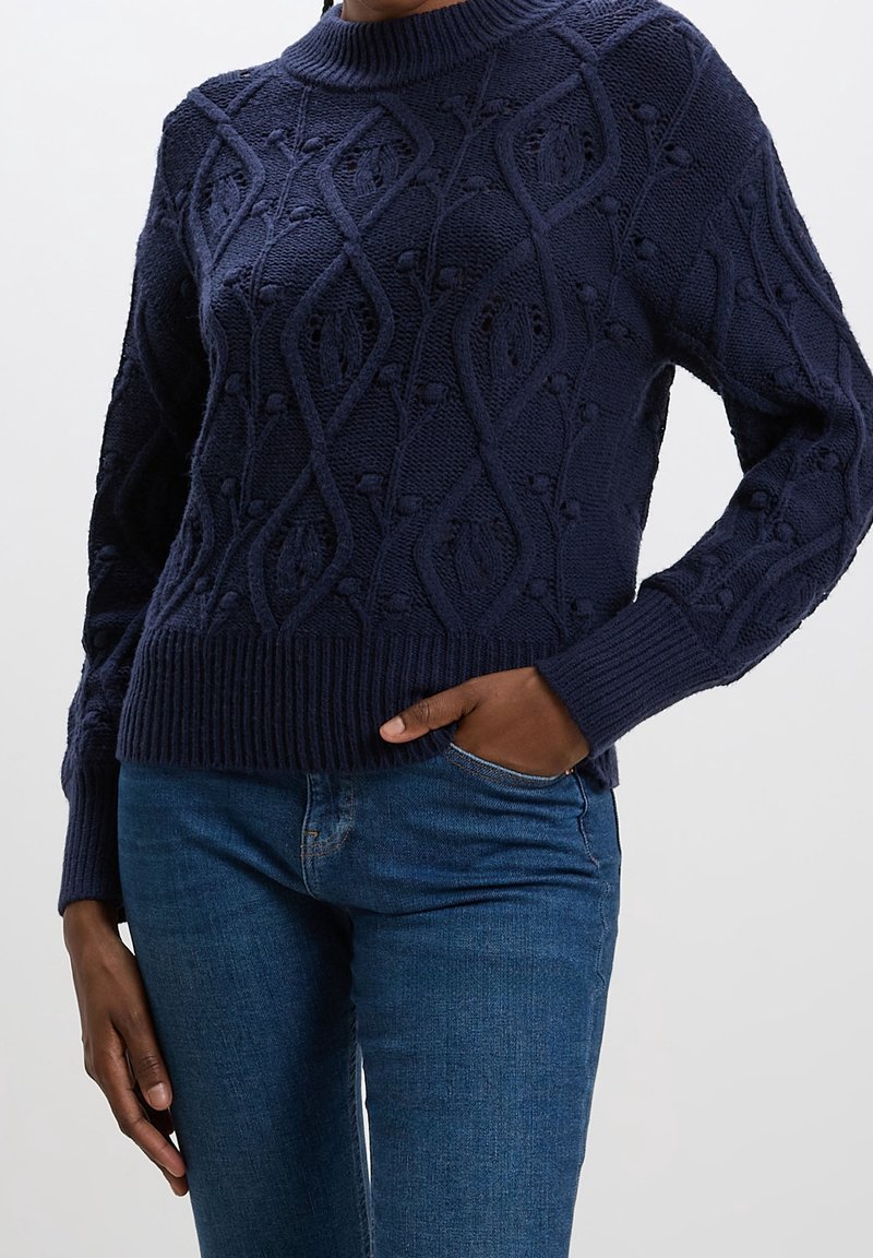 Pull torsadé bleu marine avec motif floral porté avec un jean bleu, une main dans la poche, sur un fond uni clair.
