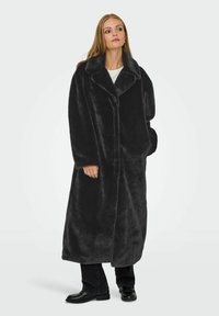Donna che indossa un lungo cappotto di pelliccia sintetica nera sopra pantaloni neri e mocassini, in piedi con una mano in tasca su uno sfondo chiaro e semplice.