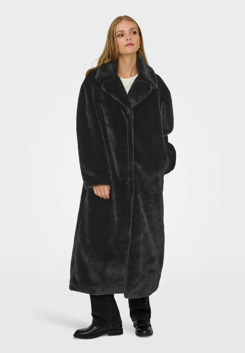 Donna che indossa un lungo cappotto di pelliccia sintetica nera sopra pantaloni neri e mocassini, in piedi con una mano in tasca su uno sfondo chiaro e semplice.