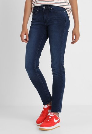 Person trägt dunkelblaue Skinny-Jeans, ein gestreiftes Hemd und leuchtend rote Nike-Sneaker mit weißen Swoosh-Logos.