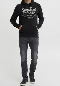 Felpa nera con cappuccio e tasca frontale, decorata con testo bianco. Abbinata a jeans neri sbiaditi e sneakers nere.