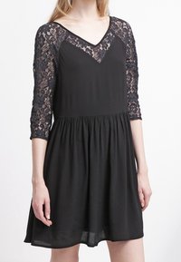 Vestido negro con mangas de encaje de tres cuartos, escote en V, corsé ajustado y falda acampanada. Tejido ligero con textura suave.