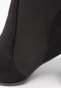 Bottine noire à la cheville avec panneau latéral élastique et petit talon bloc, montrant une texture détaillée et des coutures sur les matériaux en tissu et en daim.