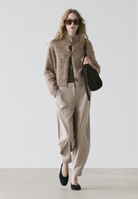Brun strikket cardigan med tekstur og cropped design, kombineret med lyse beige bukser og sorte flade sko. Tilbehør inkluderer store solbriller og en sort taske.