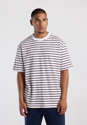 Junger Mann trägt ein weißes T-Shirt mit kurzen Ärmeln und dünnen schwarzen und roten horizontalen Streifen, steht vor einem schlichten hellen Hintergrund.