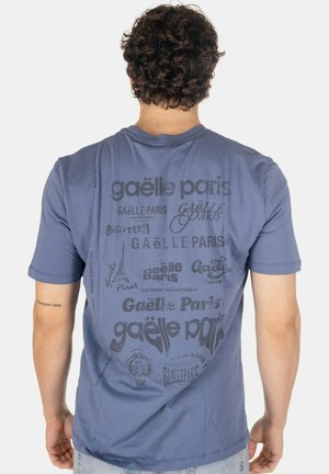 Uomo che indossa una t-shirt blu Gaëlle Paris con molteplici loghi stampati e variazioni di testo sulla schiena, in piedi davanti a uno sfondo semplice.