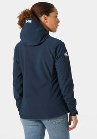 Marineblaue Softshelljacke mit einer enganliegenden Kapuze, elastischen Bündchen und einem weißen Logo auf dem Ärmel, entworfen für Komfort und Bewegungsfreiheit.