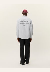 Chemise boutonnée en gris clair avec un motif à rayures verticales, comportant un texte noir au dos. Associée à un pantalon sombre et des chaussures noires.
