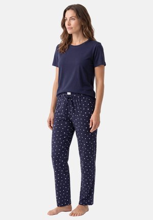Donna che indossa una maglietta blu navy a maniche corte e pantaloni da pigiama blu navy con motivo a cuori bianchi, in piedi a piedi nudi su uno sfondo semplice.