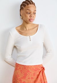 White, ribbed long-sleeve top with a scoop neckline and three buttons at the chest, paired with an orange floral wrap skirt.