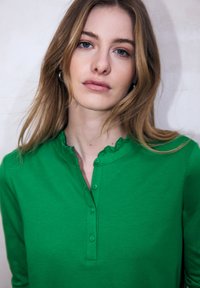 Zielona koszula z długim rękawem, z falistym kołnierzykiem, listwą z guzikami i miękką fakturą, na neutralnym tle.