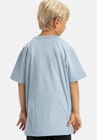 Blond dziecko stojące tyłem, noszące za dużą jasnoniebieską koszulkę i czarne spodnie na tle jednolitego białego tła.