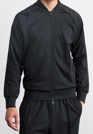 Mężczyzna ubrany w czarną sportową kurtkę Adidas na zamek oraz pasujące czarne spodnie dresowe, z rękami swobodnie opuszczonymi przy bokach.