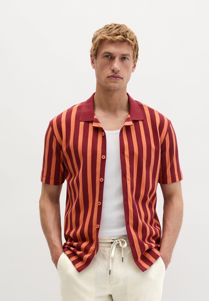 Junger Mann trägt ein kurzärmeliges Hemd mit roten und orangefarbenen vertikalen Streifen über einem weißen Tanktop und beige Zugbandhosen.