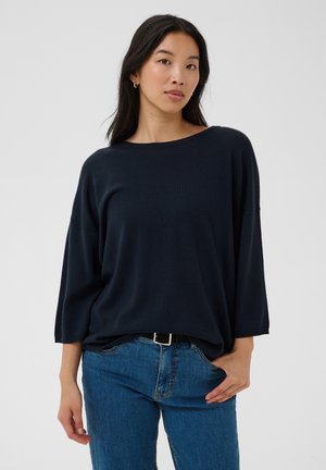 Femme aux longs cheveux foncés portant un pull bleu marine ample et un jean bleu, debout avec une main dans la poche devant un fond uni.