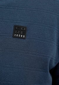 Σκούρο μπλε υφασμάτινη υφή με μαύρο τετραγωνικό έμπλαστρο που δείχνει ανάγλυφο κείμενο "CORE JACK & JONES" σε ένα ένδυμα.