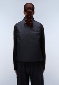 Schwarze ärmellose Steppweste, leichtes Material, hoher Kragen, glatte Textur, elastischer Saum, über einem langärmeligen braunen Oberteil getragen, Rückansicht.
