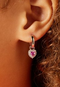 Zilveren creooloorbel met een hartvormige paarse edelsteen omringd door heldere steentjes, hangend aan een glad slotje. Krullend haar is zichtbaar.