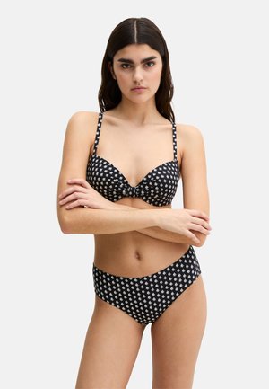 Femme aux longs cheveux foncés portant un bikini noir orné de petits motifs de feuilles blanches, debout les bras croisés sur un fond uni.